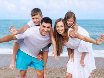 A mother and father, on holiday, pose for the camera at the shore of a beach, with their young children on their backs.