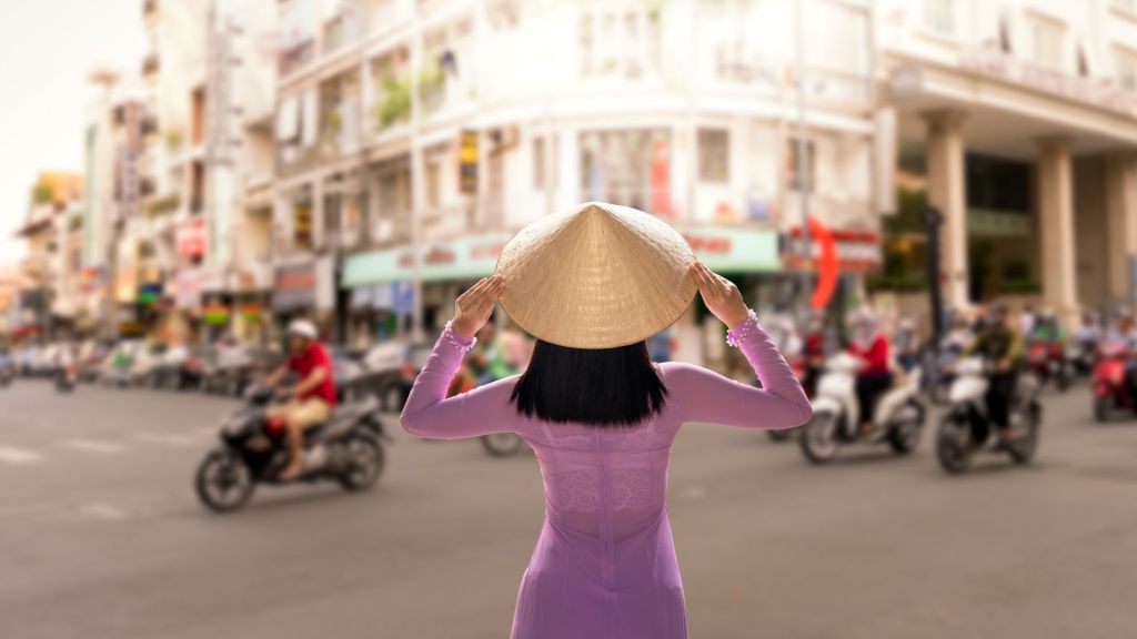 Woman in Vietnamese costume stands looking out at a street in Ho Chi Minh City, Vietnam.