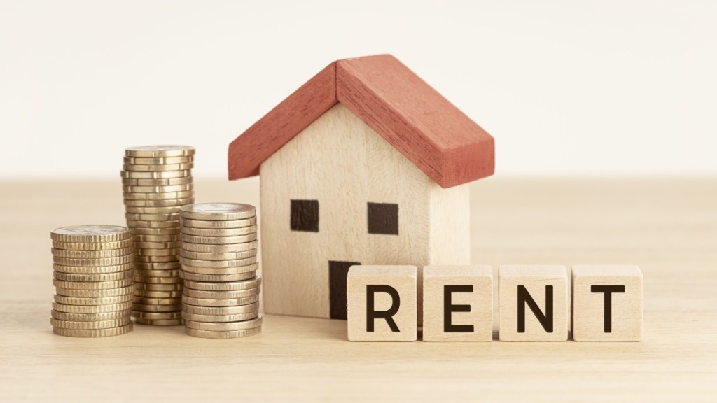The word 'RENT' spelled out on miniature wooden bricks in front of a wooden house with stacks of coins by the side.