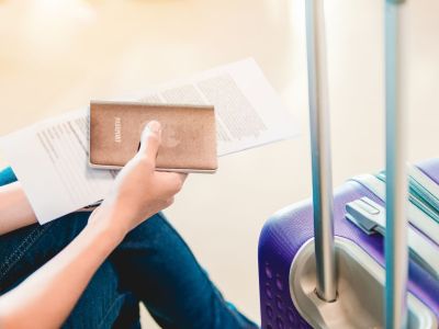 A female passenger is waiting at an airport with her suitcase beside her and her passport in her hand.