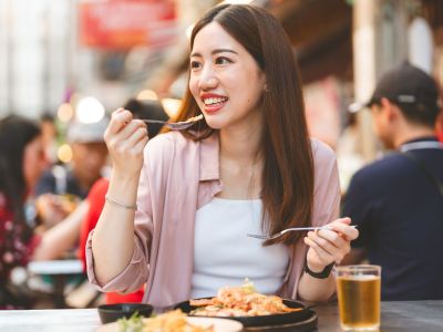 A female traveller is sitting at an outdoor table happily eating street food.