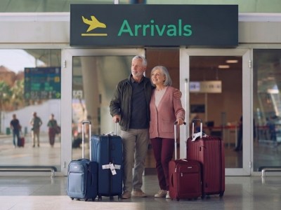 Retired couple standing with suitcases in front of the arrivals entrance of an airport.